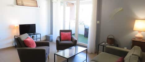 A modern living room with a flat-screen TV, two armchairs, a sofa, a glass coffee table, and a lamp.