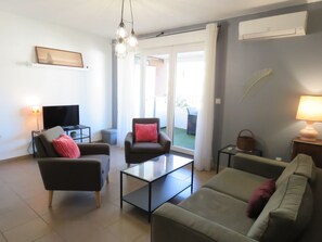 A modern living room with a flat-screen TV, two armchairs, a sofa, a glass coffee table, and a lamp.
