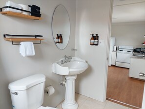 Un baño con inodoro, lavamanos y espejo. Hay estantes con toallas y botellas. Se ve una cocina a través de una puerta abierta.