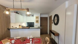 A dining area with a wooden table set for four, a kitchen in the background, and a clock on the wall. - Beautiful bright apartment in the center, with parking (Les Gets)