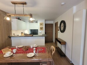 Un comedor con una mesa de madera dispuesta para cuatro, una cocina al fondo y un reloj en la pared.