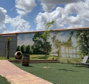 Un espacio exterior ajardinado con césped artificial, un mural de árboles, un barril de madera y una silla de descanso.