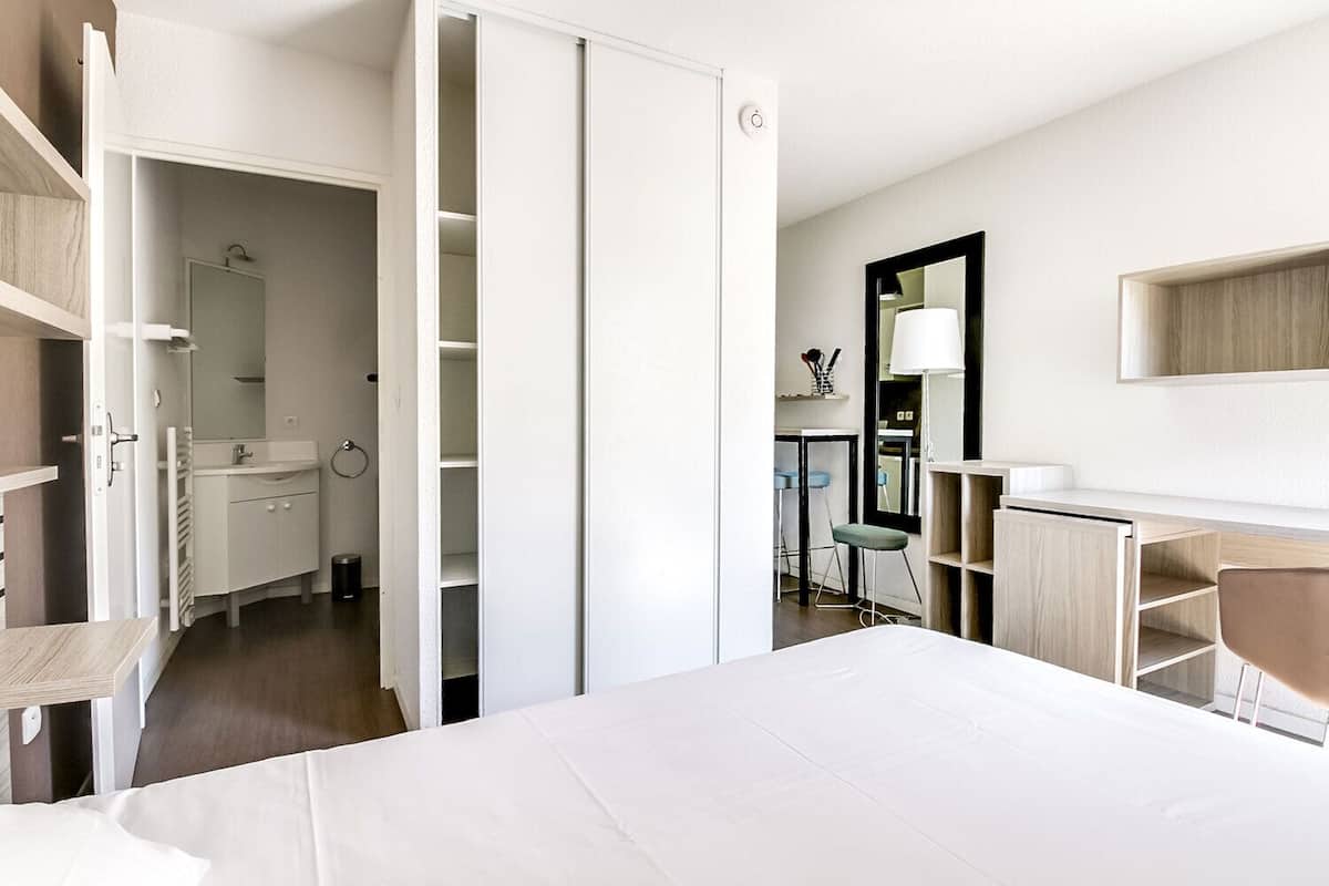 A modern hotel room with a large bed, a desk, a chair, a wardrobe, and a bathroom with a sink and mirror.