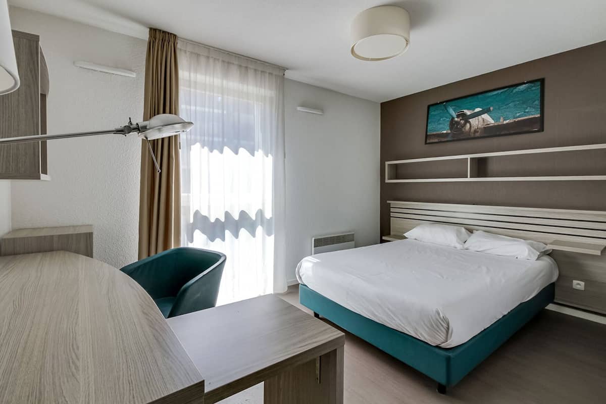 A modern hotel room with a bed, a desk, a chair, a lamp, and a painting on the wall.