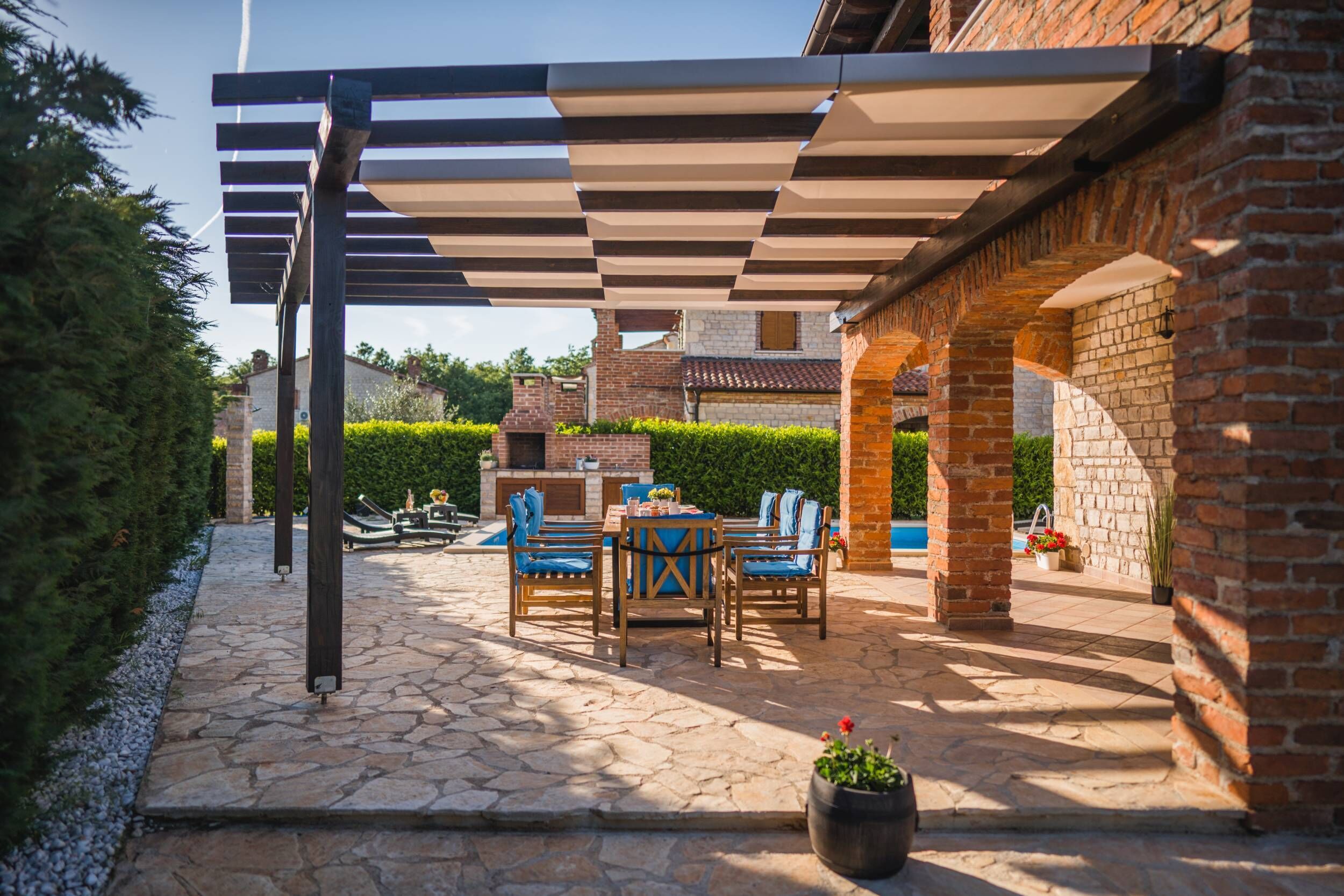 Une terrasse extérieure avec un sol en pierre, une table et des chaises en bois, et une structure en forme de pergola.