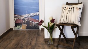 Une pièce dotée d’un plancher en bois, d’une chaise avec un coussin, d’un téléviseur à écran plat fixé au mur, et d’une porte offrant une vue panoramique sur une ville côtière et des montagnes.