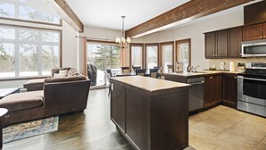 A modern kitchen with a large island, dark wooden cabinets, stainless steel appliances, and large windows offering a view of the outdoors.