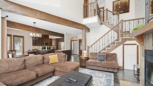 A spacious living room with a large sectional sofa, a black ottoman, and a patterned rug. A staircase with wooden railings leads to an upper level.