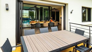 Ein Essbereich im Freien mit Holztisch und -stühlen, eine Glastür zum Pool und Blick auf das Pool-Areal.