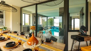 Ein Essbereich mit einem langen Tisch, gedeckt für eine Mahlzeit, einem großen Fenster mit Blick auf einen Swimmingpool und einer Statue auf einem Beistelltisch.
