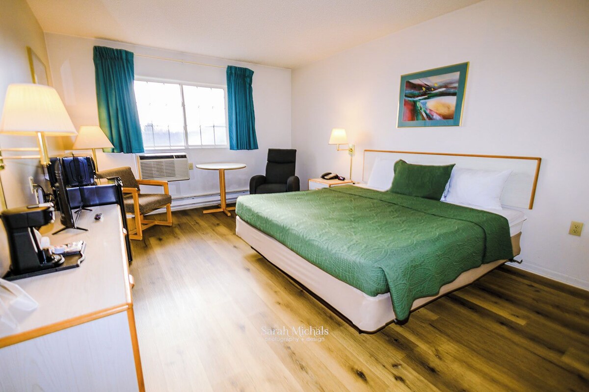 A hotel room with a bed, a desk with a lamp, a chair, a small table, and a window with curtains.