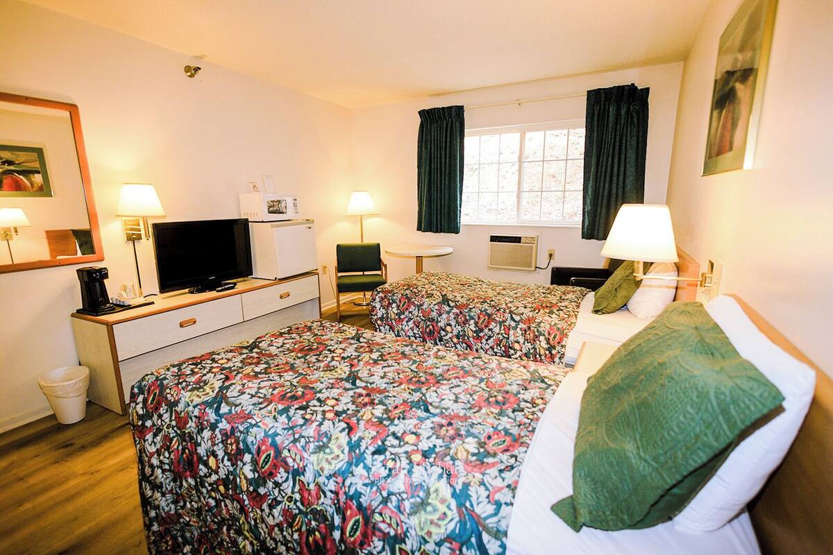 A hotel room with two beds, a desk with a TV, a small fridge, and a window with curtains.