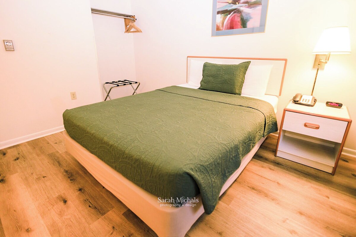 A neatly made bed with a green bedspread, a bedside table with a lamp, and a wall-mounted towel rack.