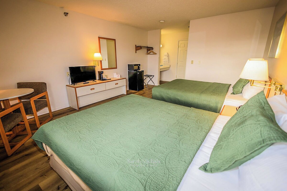 A hotel room with two beds, a desk, a chair, a dresser, a mirror, a lamp, and a microwave.