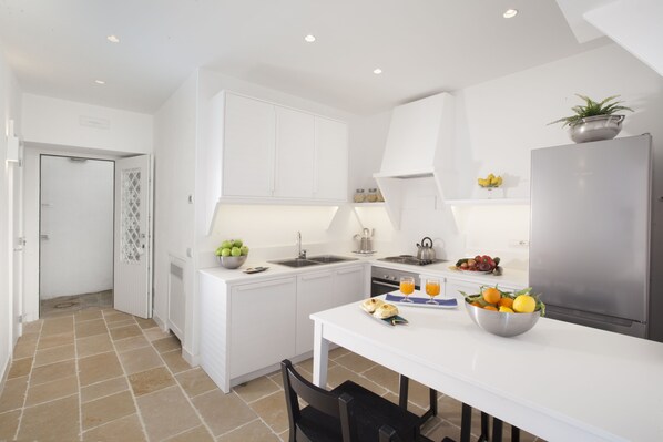 A modern kitchen with white cabinetry, a stainless steel refrigerator, and a dining table with a bowl of fruit and a kettle. - Sea Beauty (Massa Lubrense)