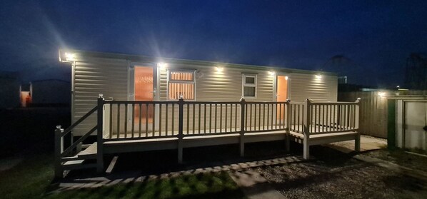 A single-story mobile home with a front porch, a small attached garage, and a well-lit exterior at night. - Staycation Opposite Fantasy Island Ingoldmells (Skegness)