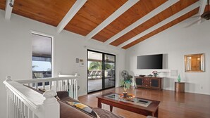 Un salon spacieux avec un plafond en bois, une grande télévision et un balcon aménagé avec du mobilier d’extérieur.