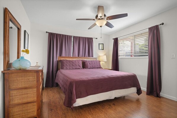 Une chambre à coucher avec un lit, un ventilateur de plafond, une commode et une fenêtre avec des rideaux.
