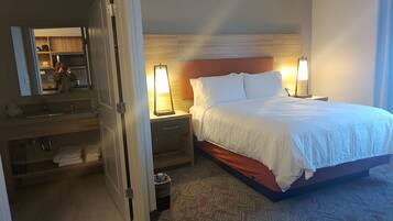Une chambre d’hôtel avec un lit, des tables de chevet, un miroir et une salle de bain visible à travers une porte ouverte.