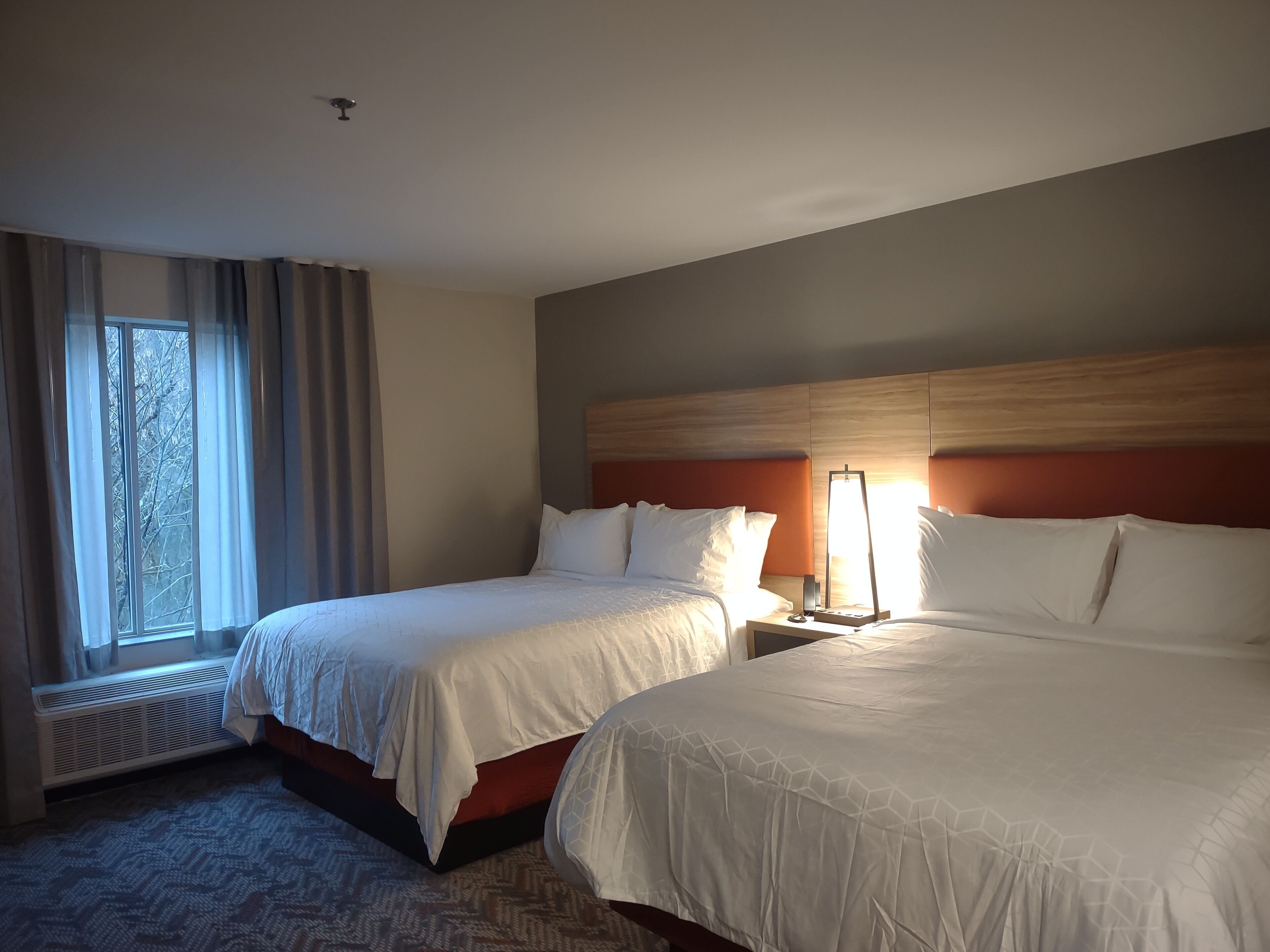 Habitación de hotel con dos camas, una ventana con cortinas, un radiador y un cabecero con una lámpara.