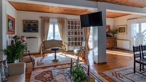 Ein geräumiges Wohnzimmer mit Holzböden, einem großen an der Wand befestigten Fernseher und Blick ins Freie durch große Fenster.