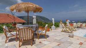 Eine Terrasse im Freien mit Korbstühlen, einem Tisch, einem Reetdachschirm und Blick auf Meer und Berge.