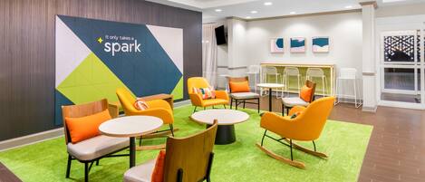Uma área de lounge moderna com design geométrico na parede, diversas opções de assento incluindo cadeiras e uma cadeira de balanço, e um tapete verde.
