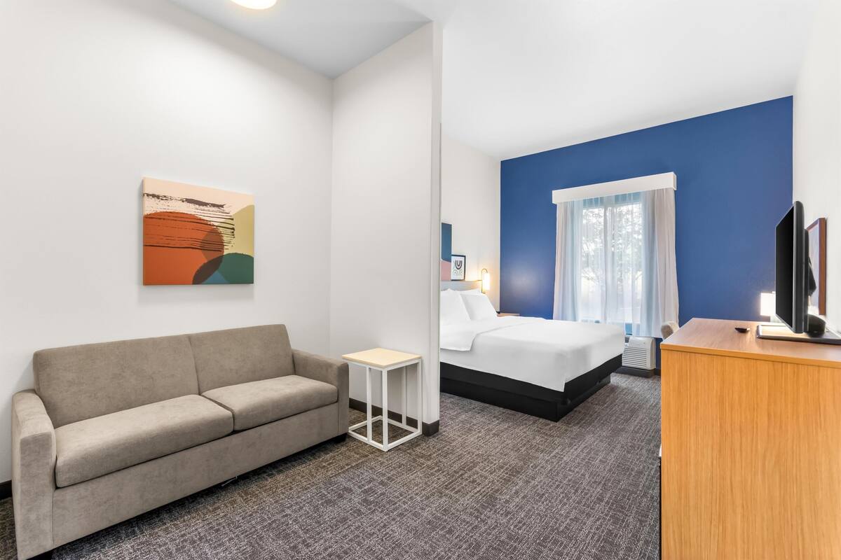 A hotel room with a sofa, a small table, a bed, a TV, and a painting on the wall.