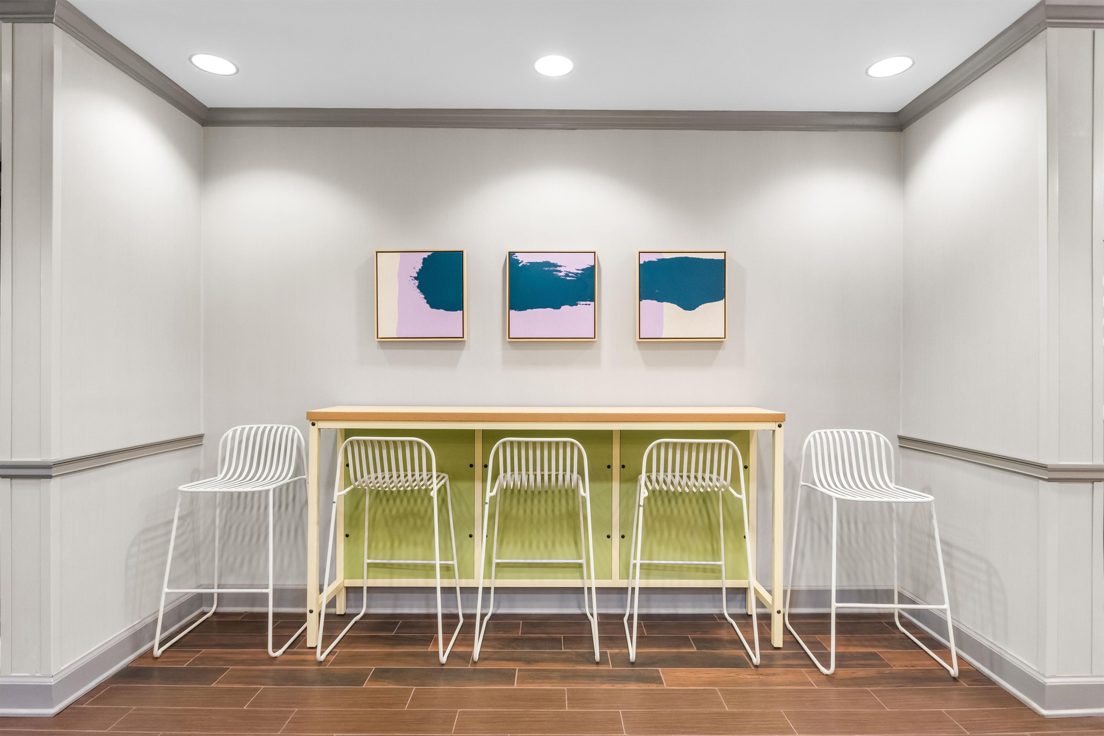 A room with a white wall, three framed artworks, a wooden counter, and four white chairs.