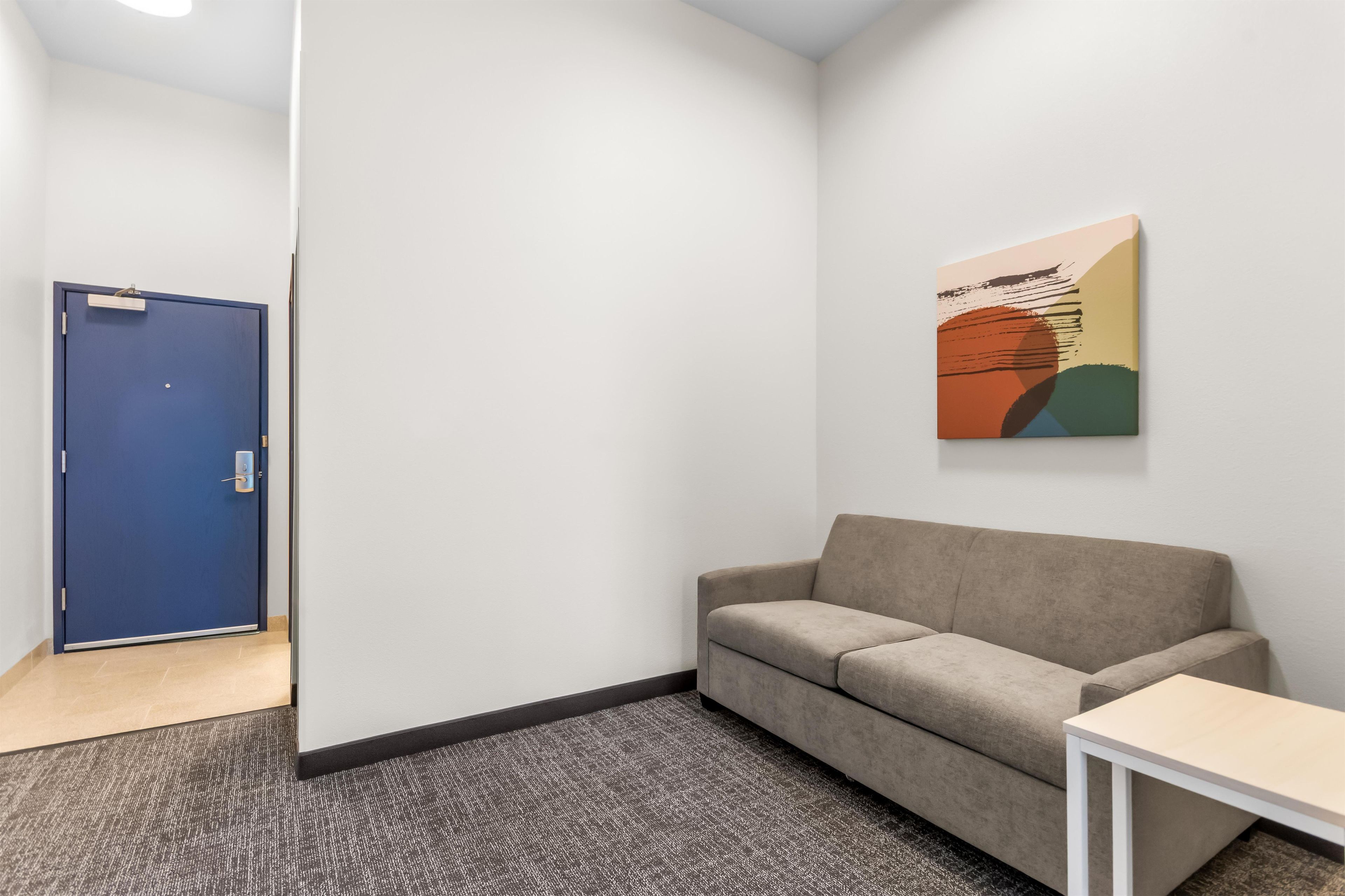 A room with a blue door, a beige sofa, a small white table, and a colorful abstract painting on the wall.
