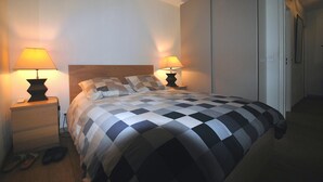 Un letto rifatto con cura, con copriletto a quadretti, due abat-jour e un comodino in legno con un paio di pantofole.