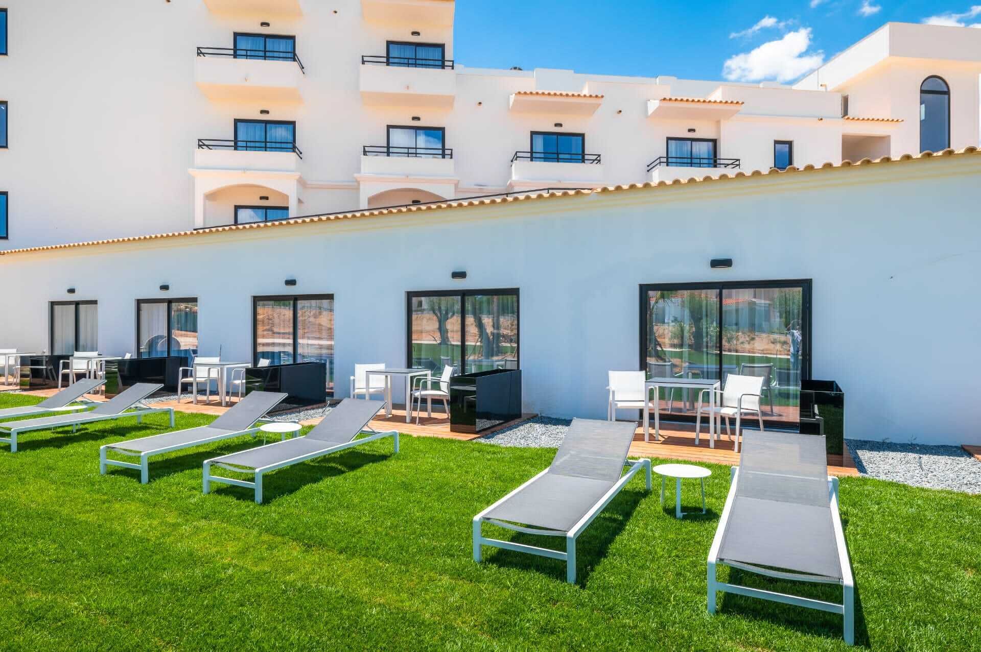 A modern outdoor lounge area with sun loungers, a barbecue, and a poolside table, overlooking a building with balconies.