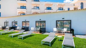 A modern outdoor lounge area with sun loungers, a barbecue, and a poolside table, overlooking a building with balconies.