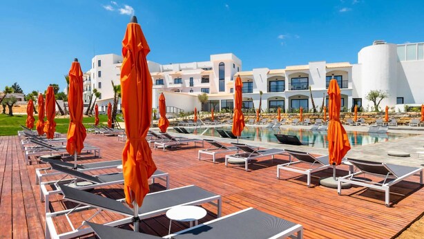 Zona de piscina en un resort con tumbonas y sombrillas naranjas, rodeada de edificios blancos de diseño moderno.