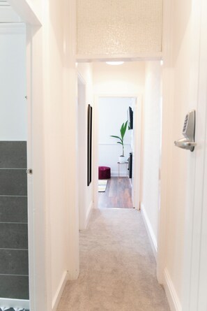 A narrow hallway with a carpeted floor leading to a room with a plant, a small table, and a red chair.