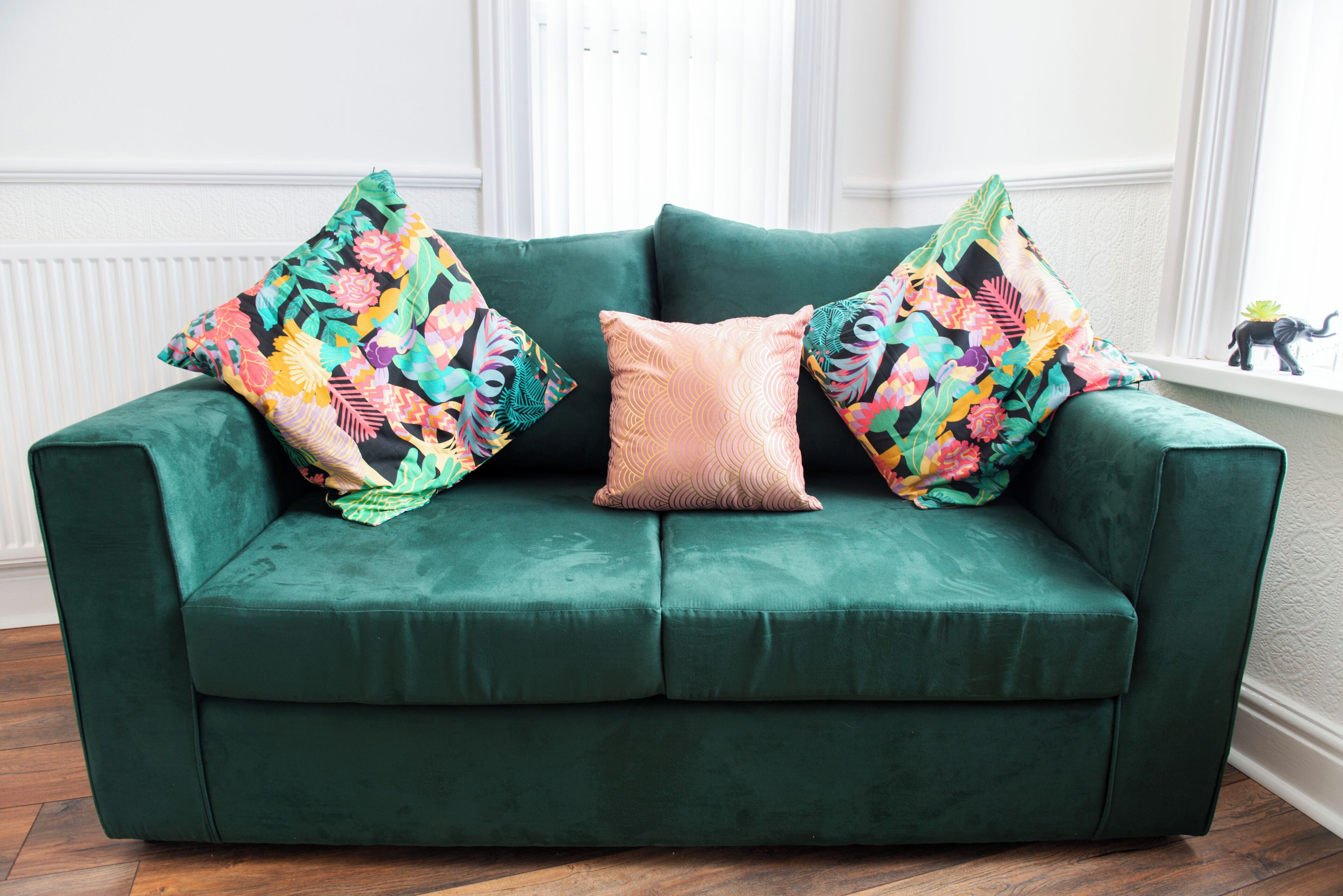 A green sofa with floral-patterned pillows and a solid-colored pillow in a room with wooden flooring and white walls.