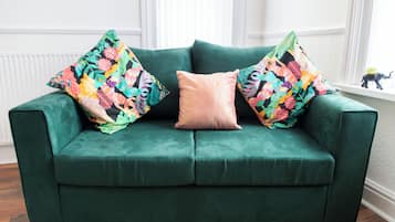 A green sofa with floral-patterned pillows and a solid-colored pillow in a room with wooden flooring and white walls.