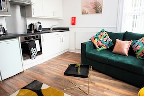 A modern kitchen with white cabinets, a black countertop, and stainless steel appliances. There is a green sofa with colorful cushions and a small black side table with a plant.