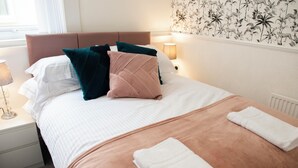 Una cama bien hecha con un cabecero marrón, almohadas blancas y color turquesa, y una manta color beige. Hay dos toallas blancas dobladas sobre la cama.