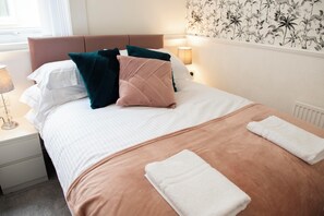 A neatly made bed with a brown headboard, white and teal pillows, and a beige blanket. There are two white towels folded on the bed. - Highcliffe Ebony Suite (Cleveleys)