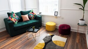 Un sofá verde con almohadones de colores, una pequeña mesita auxiliar con una planta, un otoman amarillo, un pouf rojo y una planta en maceta sobre una mesita.
