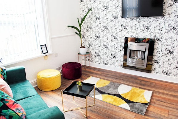 Un salon avec un canapé vert, un pouf jaune, une table basse noire, un coussin rouge, une plante en pot, une cheminée et un téléviseur fixé au mur.