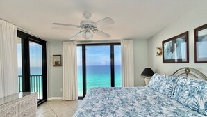 Habitación de hotel con cama, ventilador de techo, ventana grande con vista al océano y un cuadro en la pared.