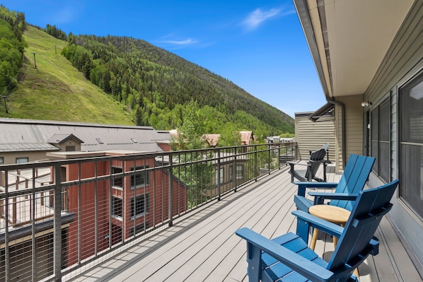 A balcony with blue chairs and a table, overlooking a forested hillside.
