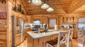 A spacious kitchen with wooden cabinets, a stainless steel refrigerator, a large island with bar stools, and a hanging light fixture. - Rocky Top Retreat  Hot Tub & Mountain Views (Suches)