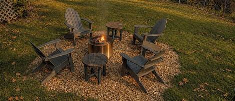 A fire pit surrounded by four Adirondack chairs and a small table, set on a gravel area with grass and trees in the background.