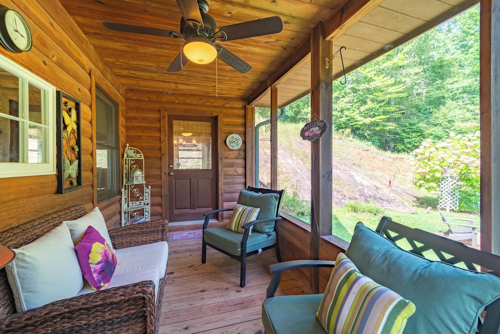 Une cabane en bois dotée d’un porche, aménagée avec un canapé et des fauteuils, un ventilateur de plafond et une porte sur laquelle est affiché un panneau d’accueil.