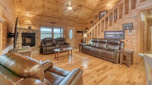 A cozy log cabin interior with a stone fireplace, leather sofas, and a wooden staircase. - Rocky Top Retreat  Hot Tub & Mountain Views (Suches)