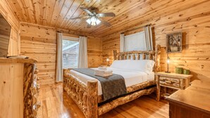 A wooden cabin bedroom with a bed, a ceiling fan, a window with curtains, and a nightstand with a lamp. - Rocky Top Retreat  Hot Tub & Mountain Views (Suches)