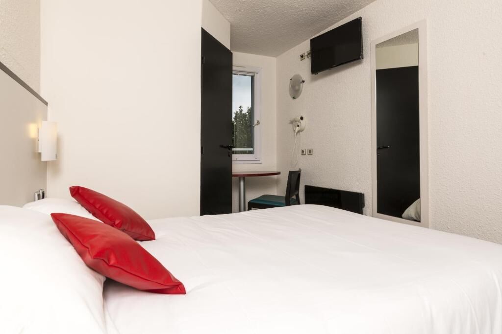 Camera d'albergo con un letto, cuscini rossi, una scrivania con una sedia, una TV a muro e una finestra con vista sugli alberi.